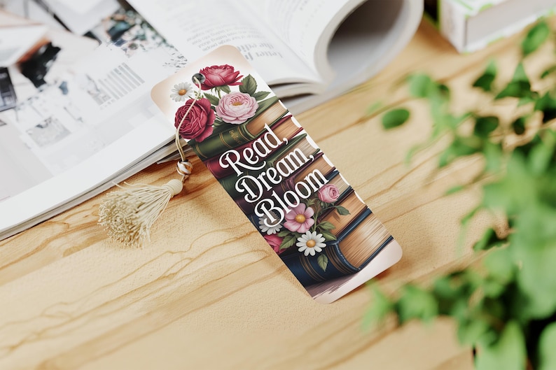 May include: A decorative bookmark featuring a floral design with red and pink roses, daisies, and the text "Read, Dream, Bloom." The bookmark has a tassel and is placed on a wooden surface next to an open book.