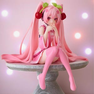 May include: A pink anime-style figurine with long, flowing pink hair and cherry-shaped accessories. The figure is wearing a pink outfit and seated on a round, stone-like table. The background is a soft pink with glowing orbs.