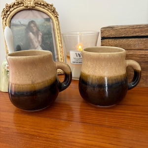 May include: Two ceramic mugs with a brown and tan glaze. The mugs have a rounded shape with a small handle. The mugs are sitting on a wooden surface. A framed picture, a candle, and a wooden box are in the background.