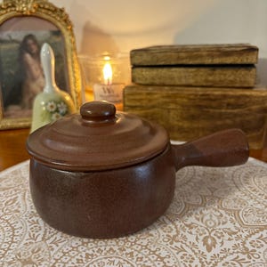 May include: A small brown ceramic pot with a lid and a handle. The pot is sitting on a white and brown patterned surface.