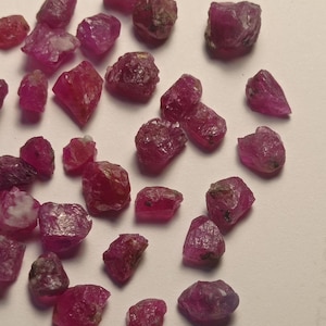 Op de afbeelding: Een verzameling ruwe, ongeslepen robijn edelstenen. De stenen variëren in grootte en vorm en vertonen een dieprode tint met enkele lichtere roze tinten. De ruwe textuur en natuurlijke vormen van de robijnen zijn zichtbaar.