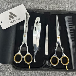 Peut inclure: Un kit de coiffeur professionnel avec un étui zippé noir. Le kit comprend des ciseaux argentés, un rasoir droit, une pince à épiler, un peigne et une étiquette avec le logo "MAXIMUS". Les ciseaux ont des trous pour les doigts dorés.