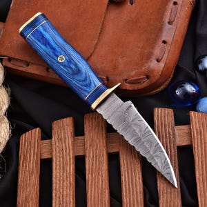 Puede incluir: Un cuchillo de hoja fija con una hoja de acero Damasco estampada y lomo dentado. El mango está hecho de madera azul con veta prominente, acentuado por virolas de latón y un pasador central de latón con una pequeña incrustación.