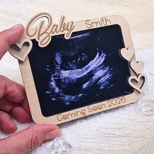 Op de afbeelding: Houten frame met een echografiebeeld. Het frame heeft het woord "Baby" in cursief, de achternaam "Smith" en de tekst "Coming Soon 2026". Decoratieve harten zijn uit het hout gesneden.