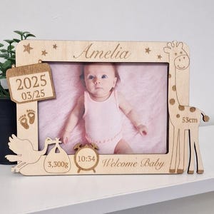 Op de afbeelding: Een houten fotolijst met een giraffen- en ooievaarontwerp. De lijst heeft een kalender, een klok en voetafdrukken. Er staat "Amelia", "2025 03/25", "10:34", "3.300 g", "53 cm" en "Welkom Baby" op.