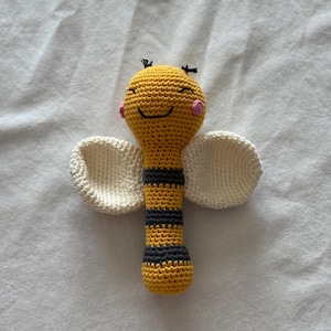 May include: A handmade crochet bee rattle. The bee has a yellow body with black stripes, white wings, and a smiling face with pink cheeks. The rattle is designed for babies and young children.