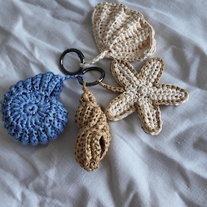 May include: A selection of crocheted keyrings with a marine theme. The set features a blue seashell, a tan seashell, a starfish, and a fan shape. Each keyring is attached to a silver-coloured ring.