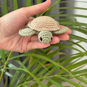 May include: A handmade crochet turtle in shades of tan and green. The turtle has a tan shell, green head and limbs, and small black eyes. The turtle is held in a hand against a background of green leaves.