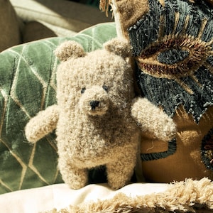 May include: A small, beige teddy bear with a textured, fuzzy appearance. The bear has a black nose and small, round ears. It is positioned in front of a green, diamond-patterned pillow and a decorative pillow with brown and blue accents.