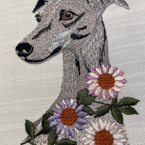Op de afbeelding: Geborduurd beeld van een windhond met een bloemenontwerp. De hond is grijs met zwarte markeringen en bruine ogen. De bloemen zijn paars, wit en roze met bruine centra en groene bladeren. De borduursels zijn op een lichtgekleurde stof.