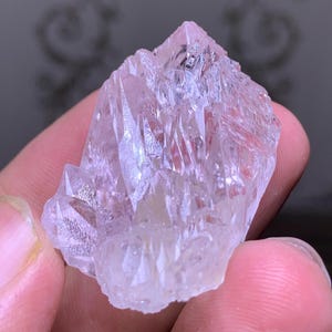 May include: A cluster of translucent, light pink quartz crystals. The crystals have sharp, pointed terminations and a rough, textured surface. The quartz is held in a hand, with the background out of focus.