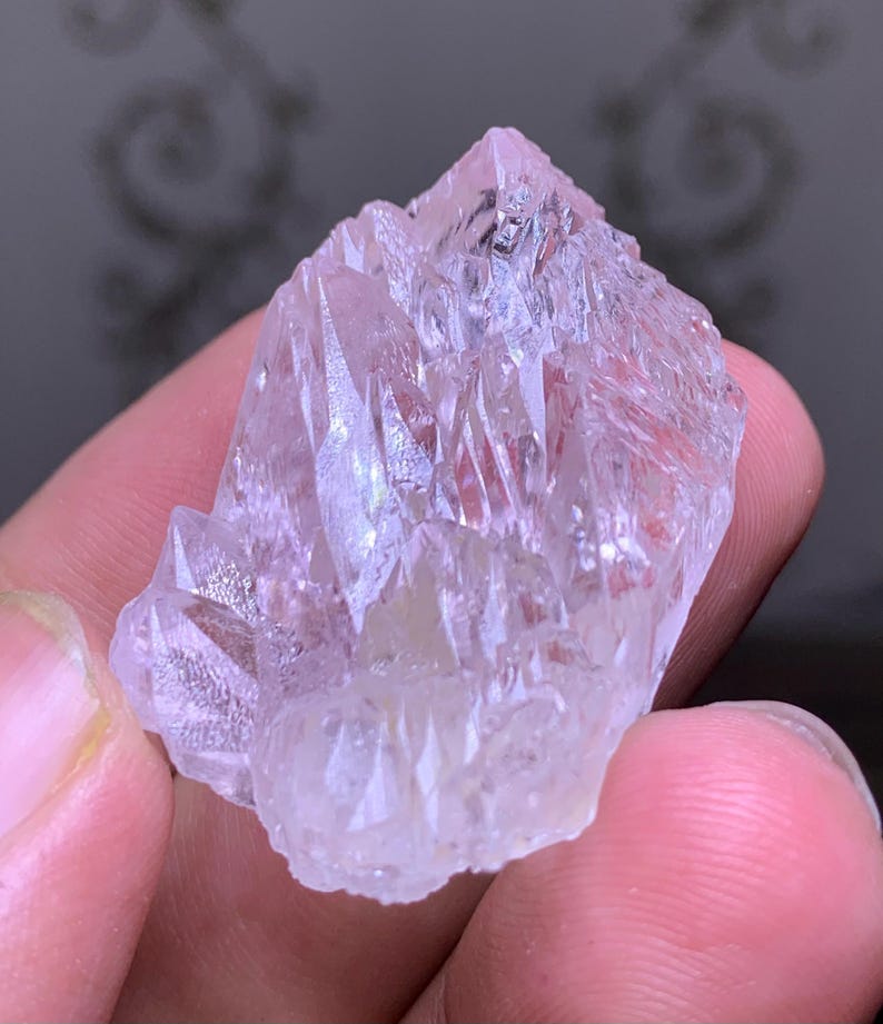 May include: A cluster of translucent, light pink quartz crystals. The crystals have sharp, pointed terminations and a rough, textured surface. The quartz is held in a hand, with the background out of focus.