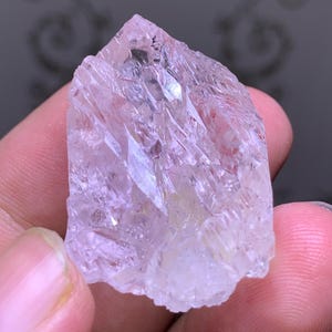 May include: A translucent, light pink crystal with a jagged, crystalline structure. The raw mineral has a rough, uneven surface and a pointed top. The crystal is held in a person's hand, showcasing its natural form and color.