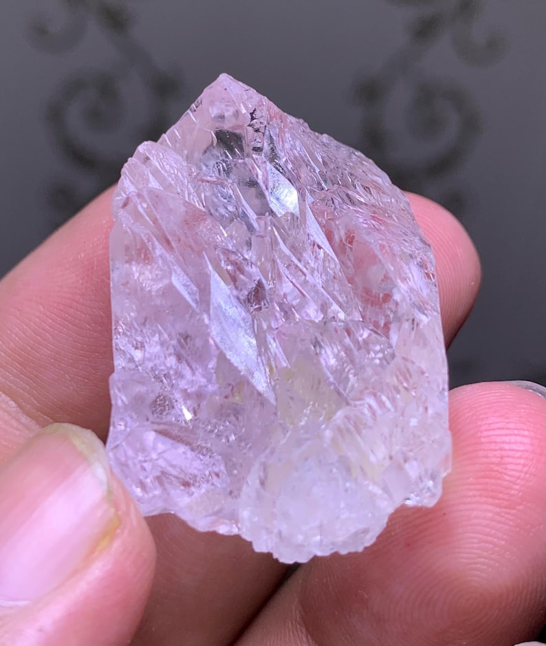 May include: A translucent, light pink crystal with a jagged, crystalline structure. The raw mineral has a rough, uneven surface and a pointed top. The crystal is held in a person's hand, showcasing its natural form and color.