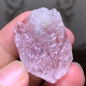 May include: A raw, uncut crystal with a pale pink hue and a rough, textured surface. The crystal has a lighter, almost white, area at the top. The facets of the crystal catch the light, creating a sparkling effect. This natural gemstone is a unique piece.