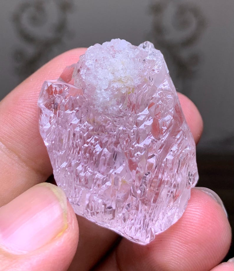 May include: A raw, uncut crystal with a pale pink hue and a rough, textured surface. The crystal has a lighter, almost white, area at the top. The facets of the crystal catch the light, creating a sparkling effect. This natural gemstone is a unique piece.