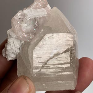 Puede incluir: Un espécimen mineral cristalino de color claro. El cuerpo principal es un cristal grande y facetado con una apariencia ligeramente translúcida. Un cristal más pequeño, rosado, se asienta sobre una base blanca y texturizada. El espécimen se sostiene en una mano.