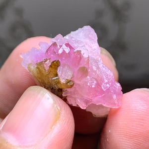May include: A cluster of pink crystals with a translucent appearance. The crystals have a vibrant, saturated pink color, with some areas appearing more opaque. The base of the cluster has a brownish-green hue.