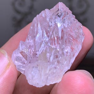 May include: A translucent, light pink crystal cluster with sharp, pointed formations. The crystal has a slightly rough texture and appears to be held in a hand. The overall shape is irregular, with multiple facets catching the light.