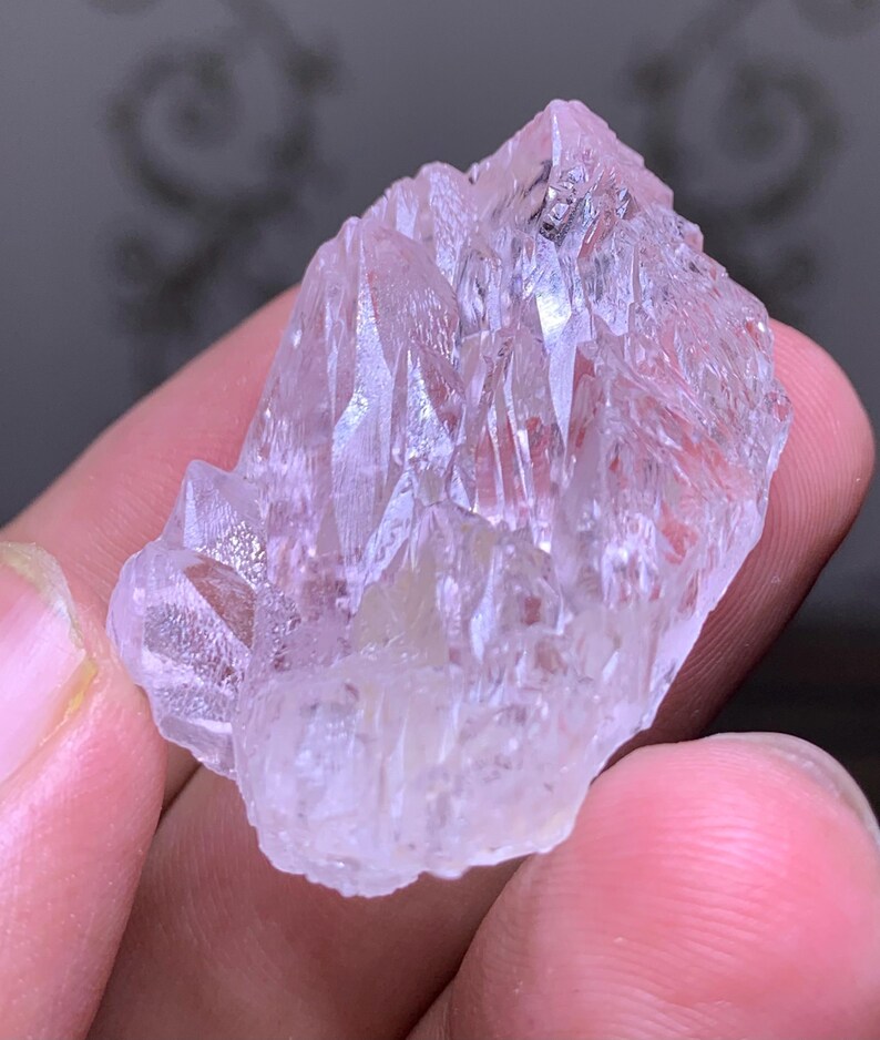 May include: A translucent, light pink crystal cluster with sharp, pointed formations. The crystal has a slightly rough texture and appears to be held in a hand. The overall shape is irregular, with multiple facets catching the light.