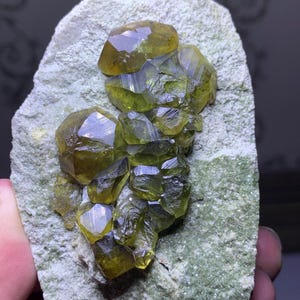 Op de afbeelding: Een cluster van doorschijnende, olijfgroene kristallen op een lichtgroene, getextureerde rotsmatrix. De kristallen hebben een facetvormige, geometrische vorm, die het licht vangt en een levendige, geelgroene tint vertoont. De rots wordt door een hand vastgehouden.