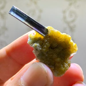 May include: A mineral specimen showing a dark, elongated crystal with blue, purple, and green colours, emerging from a cluster of yellow and green crystalline material. The mineral is held in a hand.