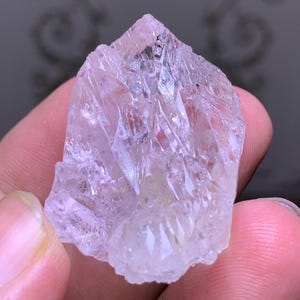 May include: A clear, light pink crystal with a pointed top and multiple facets. The crystal is held in a hand, showcasing its natural, raw form. The crystal's surface reflects light, highlighting its crystalline structure.