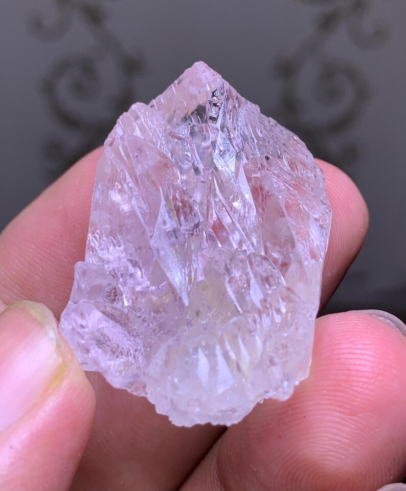 May include: A clear, light pink crystal with a pointed top and multiple facets. The crystal is held in a hand, showcasing its natural, raw form. The crystal's surface reflects light, highlighting its crystalline structure.