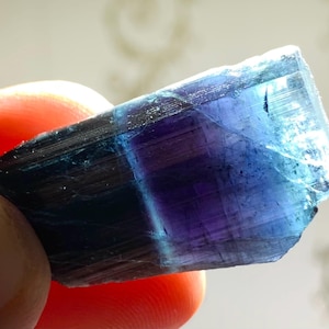 May include: A blue and purple gemstone with a striated pattern. The gemstone is rough and has a natural, unpolished look.