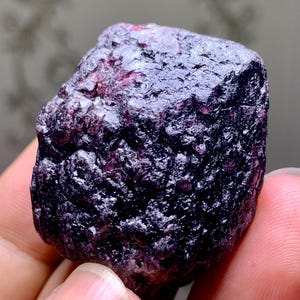 Puede incluir: Una gema rugosa de color púrpura oscuro con una superficie texturizada. La piedra tiene un color profundo con toques de rojo y una forma ligeramente irregular. Este espécimen mineral natural es ideal para coleccionistas.