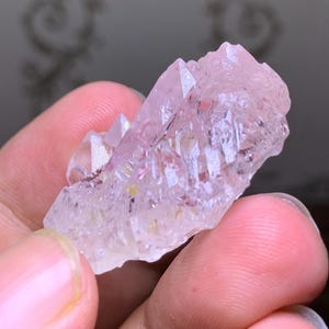 May include: A cluster of light pink quartz crystals with sharp, pointed terminations. The crystals have a translucent appearance with subtle internal color variations. The quartz is held in a hand, showcasing its natural form and texture.