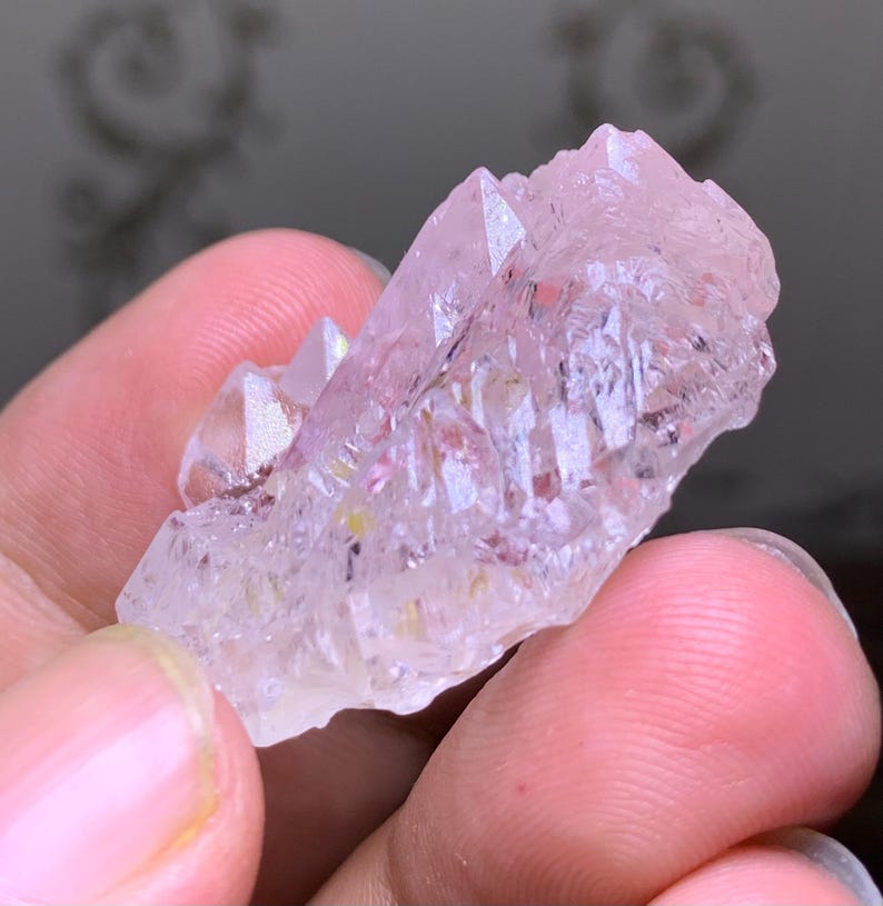 May include: A cluster of light pink quartz crystals with sharp, pointed terminations. The crystals have a translucent appearance with subtle internal color variations. The quartz is held in a hand, showcasing its natural form and texture.