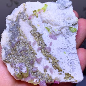 Może przedstawiać: Naturalny okaz minerału z białą matrycą i osadzonymi kryształami. Kryształy prezentują różnorodne kolory, w tym odcienie zieleni, różu i szarości. Ogólna tekstura jest szorstka i nierówna, ukazując surowe piękno minerału.