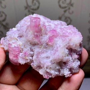 Puede incluir: Un grupo de cristales de turmalina rosa, con una prominente formación cristalina vertical. Los cristales están engastados en una matriz de minerales blancos y rosa pálido. La apariencia general es áspera y natural, mostrando la belleza en bruto de la gema.