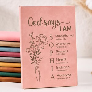May include: A stack of notebooks in various colours, with a pink cover displaying the text "God says I AM" and floral artwork. The text lists attributes such as Strengthened, Overcome, Peaceful, Heard, Included, and Accepted. Other notebooks are in pink, tan, blue, red, grey, yellow, green, and brown.