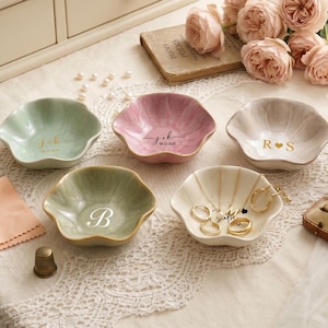 Könnte beinhalten: Fünf gewellte Keramikschalen in Pastelltönen wie Salbeigrün, Altrosa und Creme. Die Schalen sind mit personalisierten goldenen oder weißen Schriftzügen versehen, darunter Initialen wie J+K, R+S und B sowie Datumsangaben. Schmuck liegt in einer der Schalen.