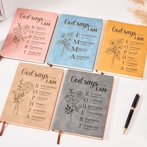 Könnte beinhalten: Fünf Notizbücher in verschiedenen Farben, darunter Rosa, Blau und Beige, mit dem Text "God says I AM" und floralen Illustrationen. Die Bücher haben eine lederähnliche Textur und ein Leseband.