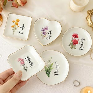 Könnte beinhalten: Eine Sammlung kleiner, weißer Keramik-Schmuckschalen in verschiedenen Formen, jede mit einem goldenen Rand und einem Blumendesign. Die Schalen sind mit Namen wie Evelyn, Sophia und Hedwig personalisiert. Eine Hand hält eine Schale.