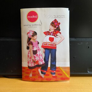 Pode incluir: Um livreto de padrões de costura para um vestido e uma camisa estilo túnica com uma bandana. A capa apresenta uma ilustração colorida de duas meninas vestindo as peças. O título do padrão é "Frida" e o texto "Mod Threads for the Cool Kid" está na parte inferior.