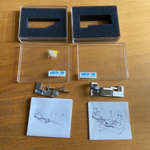 Może przedstawiać: Dwa stopki do maszyny do szycia z instrukcjami i opakowaniem. Stopki są srebrne i mają czarną plastikową podstawę. Opakowanie jest z przezroczystego plastiku z czarną wkładką z pianki. Instrukcje są wydrukowane na białym papierze i pokazują, jak używać stopek.