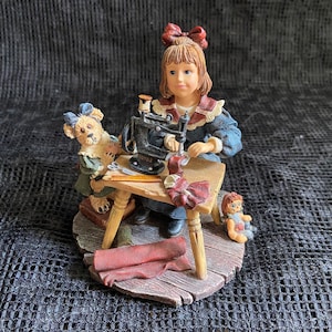 Op de afbeelding: Een beeldje van een meisje dat naait aan een tafel met een vintage naaimachine. Het meisje draagt een blauwe jurk en heeft een rode strik in haar haar. Een teddybeer en een pop zijn ook aanwezig. De basis is van hout.