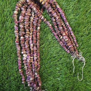Könnte beinhalten: Mehrere Stränge roher, ungeschliffener Rubin-Edelsteinperlen in Pink-, Lila- und Brauntönen. Die ungeschnittenen Perlen sind mit silberfarbenen Verschlüssen verbunden und liegen auf grünem Gras.
