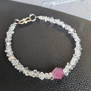 May include: A delicate bracelet featuring clear, irregularly shaped beads strung together. A central, hexagonal, deep pink gemstone adds a pop of color. The bracelet is secured with a silver-toned clasp, showcasing a blend of natural and refined elements.