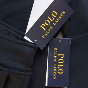 May include: Close-up of black fabric with two Polo Ralph Lauren tags. The tags are black with gold lettering and a white tag attached. The fabric appears to be a garment.