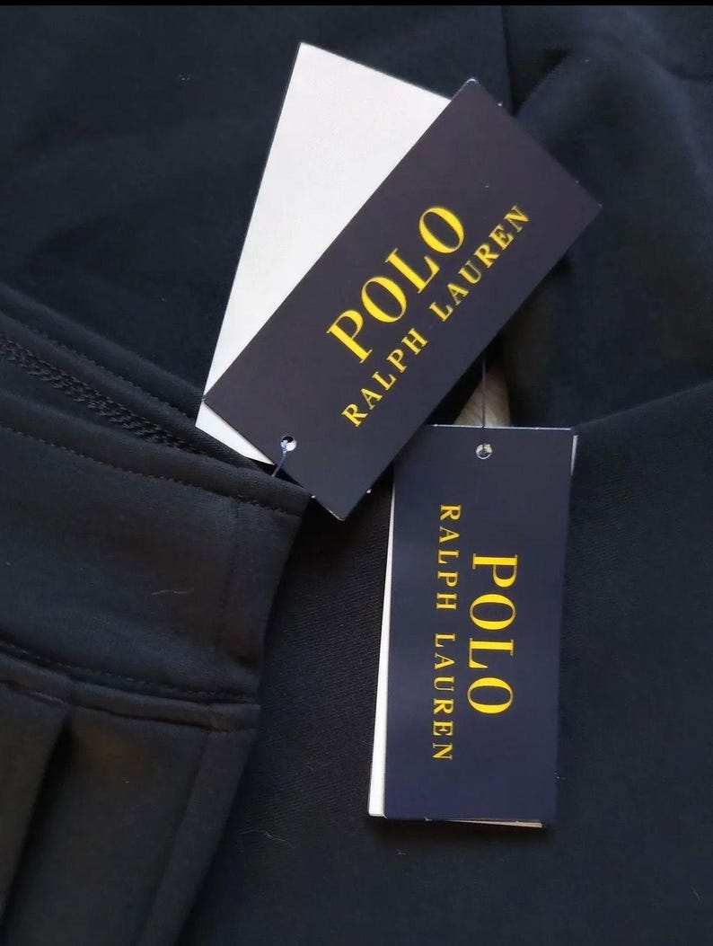 May include: Close-up of black fabric with two Polo Ralph Lauren tags. The tags are black with gold lettering and a white tag attached. The fabric appears to be a garment.