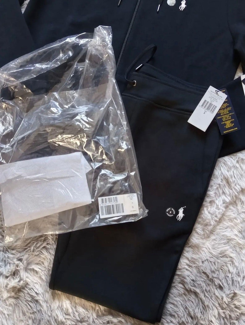 May include: Black tracksuit set, including a zip-up hoodie and sweatpants. The hoodie features a small logo on the chest, and the sweatpants have a matching logo. The set is new, with tags still attached and packaged in plastic.
