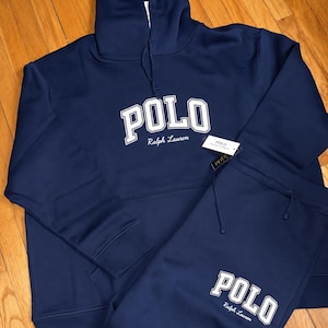 Może przedstawiać: Granatowy zestaw dresowy Polo Ralph Lauren. Słowo "POLO" jest wydrukowane białymi literami na przodzie bluzy z kapturem i spodni dresowych. Logo Ralph Lauren jest wydrukowane poniżej.