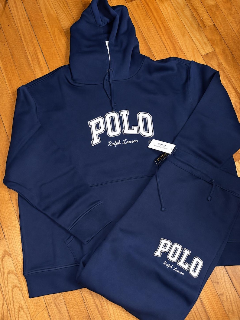 Może przedstawiać: Granatowy zestaw dresowy Polo Ralph Lauren. Słowo "POLO" jest wydrukowane białymi literami na przodzie bluzy z kapturem i spodni dresowych. Logo Ralph Lauren jest wydrukowane poniżej.