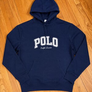 Może przedstawiać: Granatowa bluza z kapturem z napisem "POLO" dużymi białymi literami na piersi. Poniżej znajduje się tekst "Ralph Lauren". Bluza posiada kieszeń typu kangur i kaptur ze sznurkiem. Klasyczny, swobodny element garderoby.