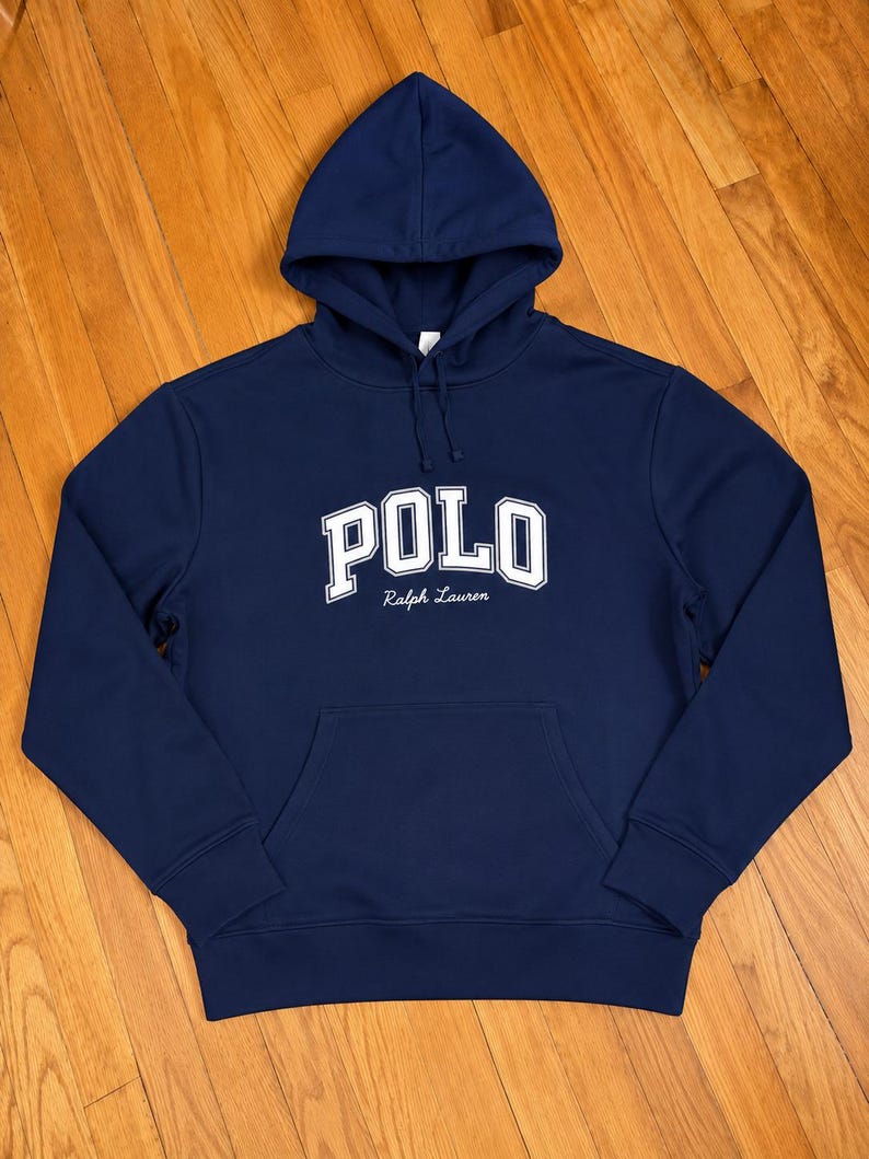Może przedstawiać: Granatowa bluza z kapturem z napisem "POLO" dużymi białymi literami na piersi. Poniżej znajduje się tekst "Ralph Lauren". Bluza posiada kieszeń typu kangur i kaptur ze sznurkiem. Klasyczny, swobodny element garderoby.